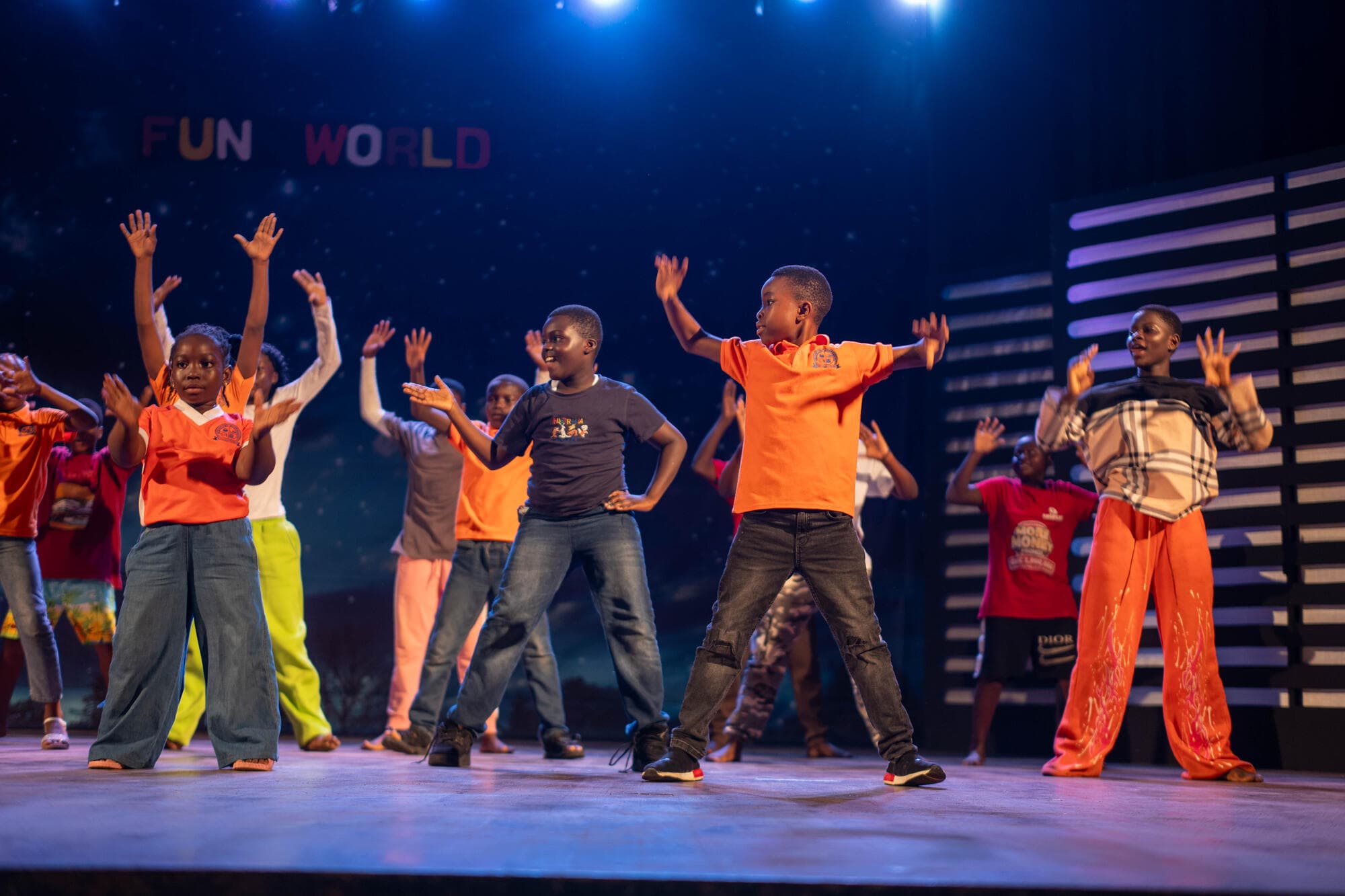 Graceland pupils performing a dance routine on the National Theatre stage under blue stage lighting, with a 'FUN WORLD' sign in the background