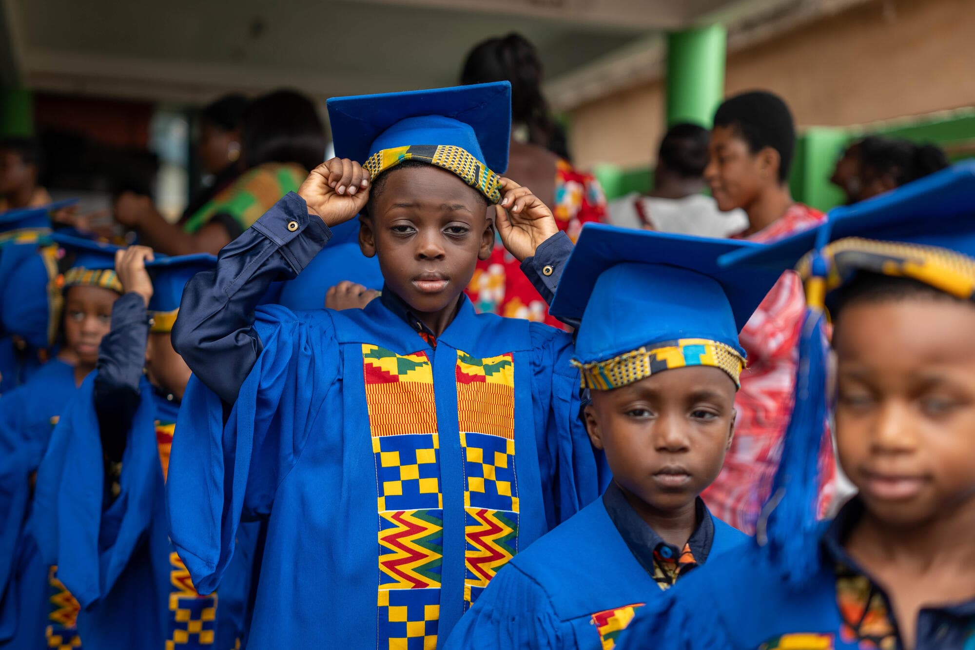 A graduating pupil in a blue cap and gown with a kente stole, adjusting his graduation cap