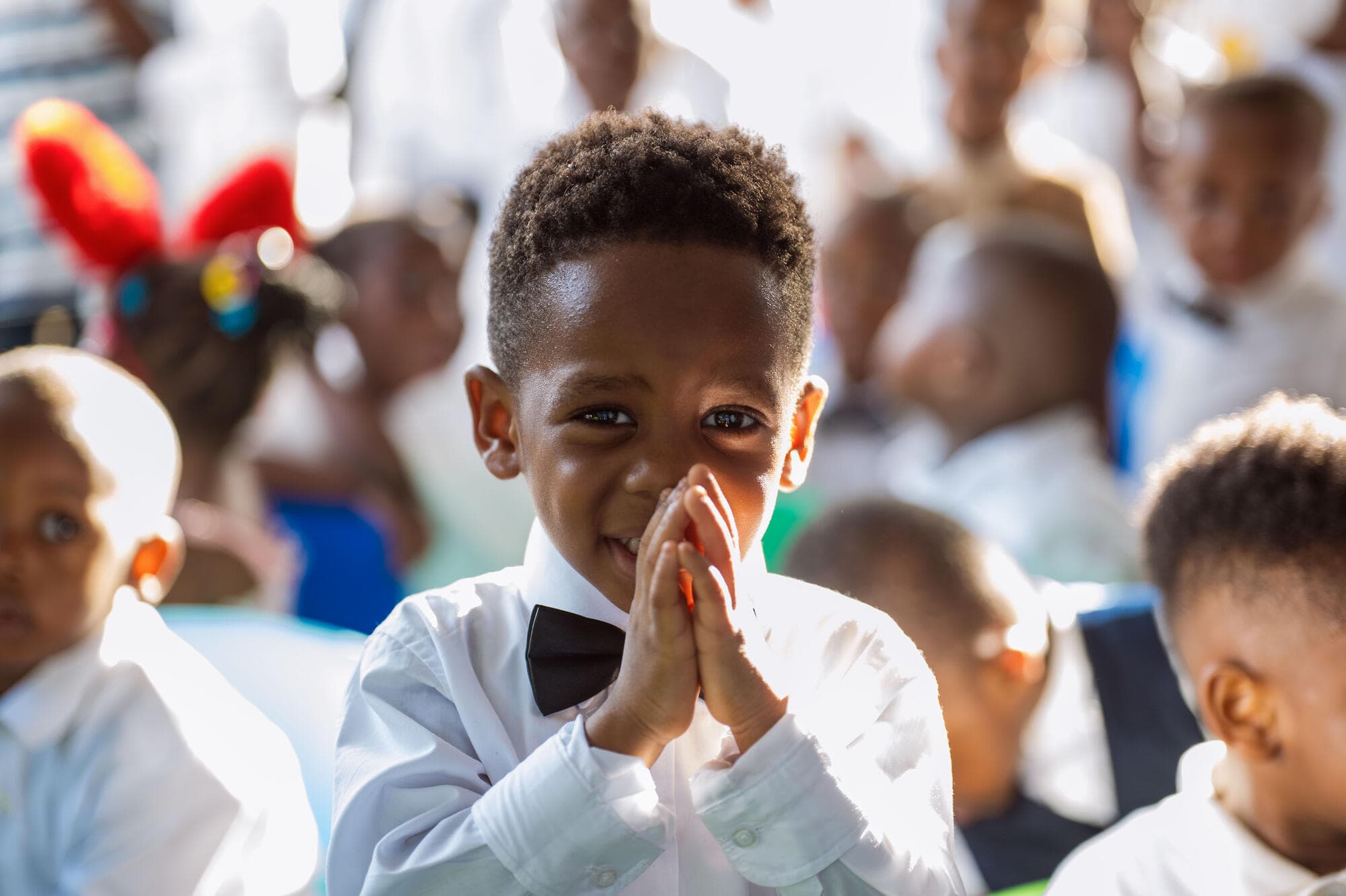 Close-up of a young pupil in a white outfit and bow tie, smiling with hands clasped together
